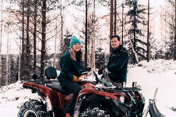 I en snöig skog sitter en leende ung kvinna på en fyrhjuling, en leende ung man står bredvid henne.